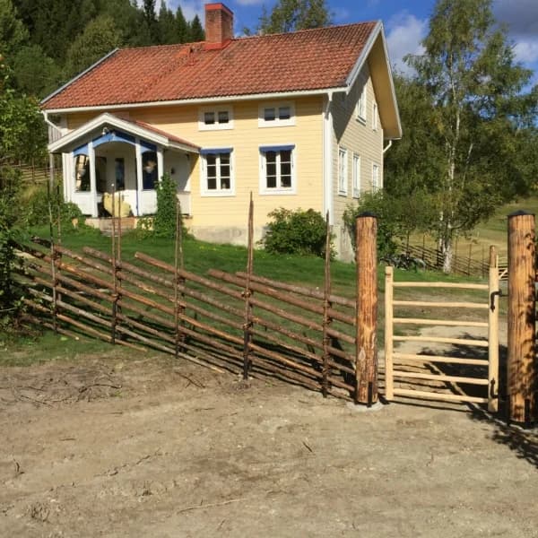 Traditionell gärdesgård med pinngrind framför ett gulmålat trähus i lantlig miljö, omgiven av grönska och skog.