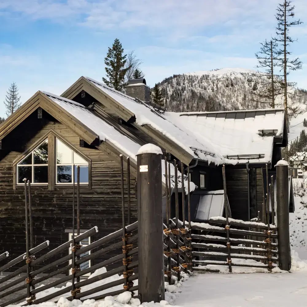 En modern, svartbetsad fjällstuga med snötäckt sadeltak och stora glasfönster. Framför huset löper en traditionell, svarttjärad gärdsgård över snöig mark.