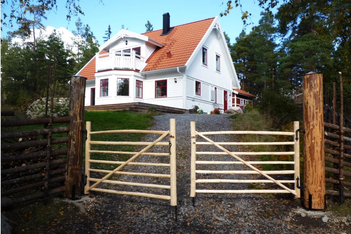 Dubbel pinngrind och traditionell gärdesgård framför ett vitmålat hus med rött tegeltak i naturnära miljö.