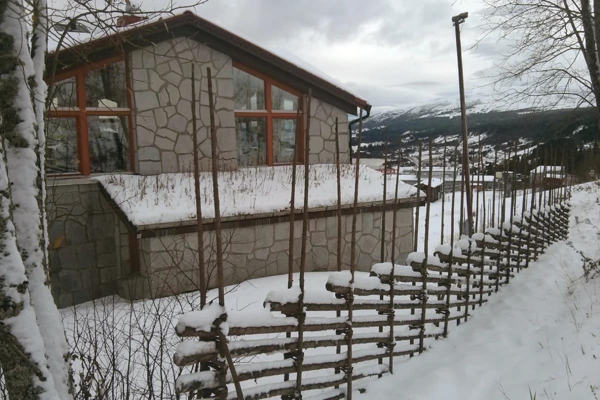 Traditionell gärdesgård täckt av snö intill ett stenklätt hus med utsikt över fjäll och vinterlandskap i Åre.