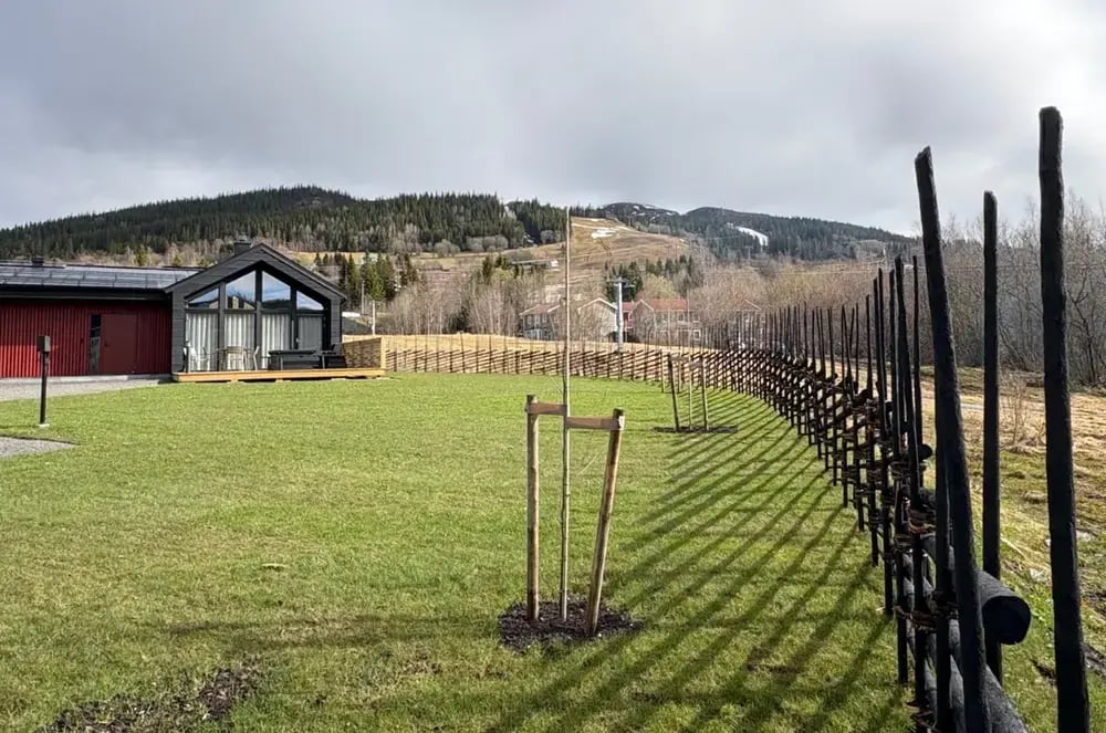 Modernt svart trähus med stora fönster, grön gräsmatta och traditionell gärdesgård med utsikt över fjäll och skidbackar i Jämtland.