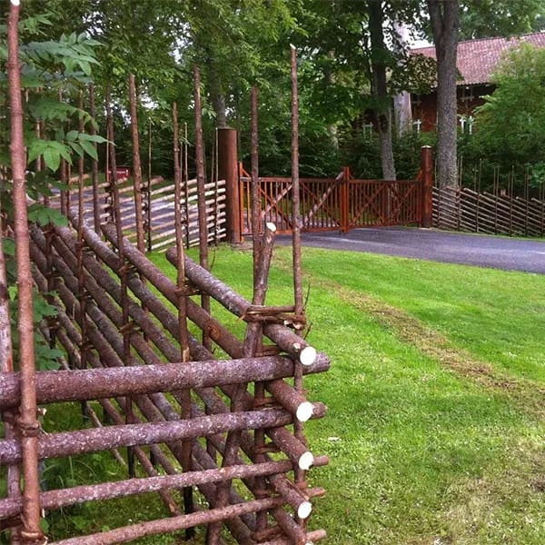 Traditionell gärdesgård med trägrind längs en grön trädgård och infart, omgiven av träd i lugn villamiljö.