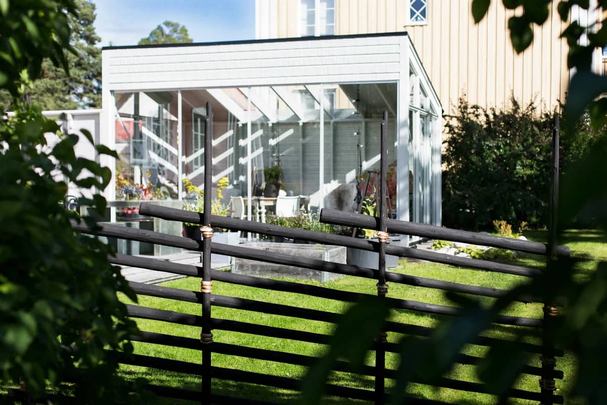 Svarttjärad traditionell gärdesgård framför ett modernt hus med inglasat uterum och grön trädgård.