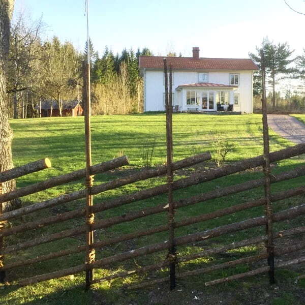 Traditionell gärdesgård framför ett vitmålat hus med stor gräsmatta, omgiven av skog och lantlig miljö.